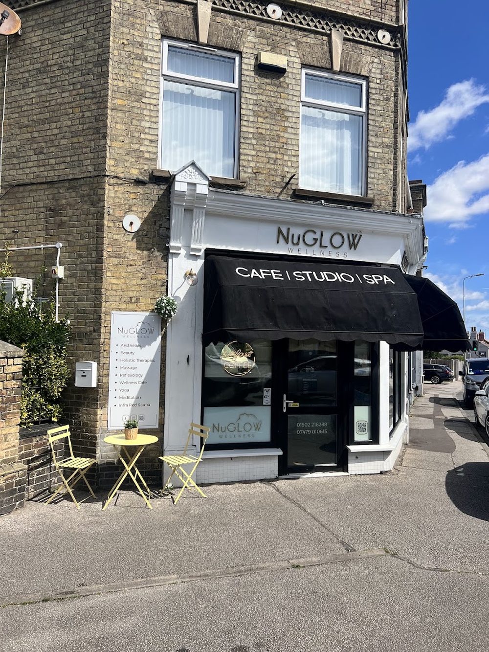 The NuGlow Wellness corner shopfront on London Road, Pakefield, with a black awning reading Cafe, Studio, Spa and bistro tables outside.